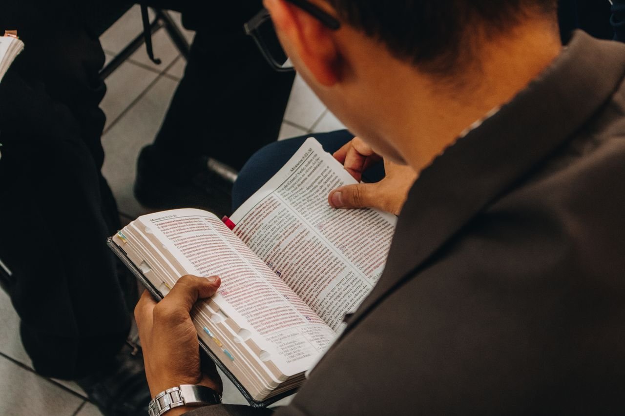 High angle back view of crop unrecognizable male in casual wear and eyeglasses sitting on bench and reading book in public place